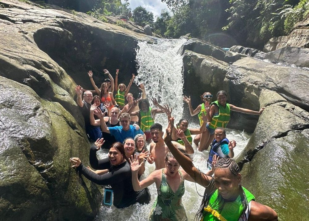 Group of people in colorful attire enjoying a waterfall surrounded by rocks and greenery.