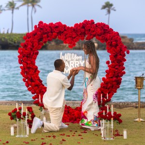 Man kneels proposing to woman in front of heart-shaped rose display by the ocean.