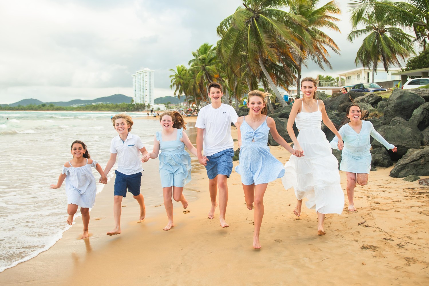 Group of seven people in beach attire running on a sandy beach near palm trees.