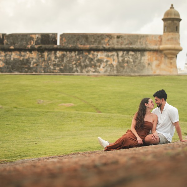 Couple sitting on grass near an old stone fortress.