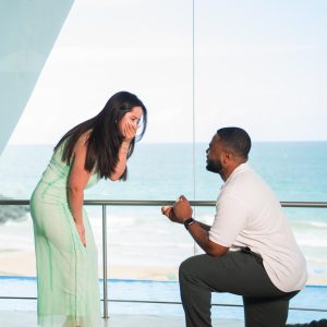 Man kneeling to propose to surprised woman by the sea indoors.
