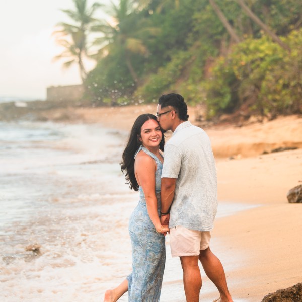 Couple standing on sandy beach with palm trees, man kissing woman's forehead.