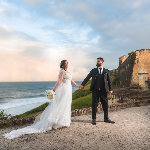 Bride and groom hold hands near an ancient stone structure by the sea.