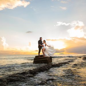 Couple in wedding attire standing on rocks by the ocean at sunset.
