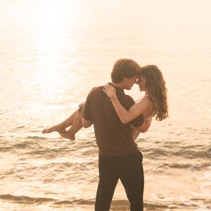 Couple embracing in ocean at sunset, man carrying woman.