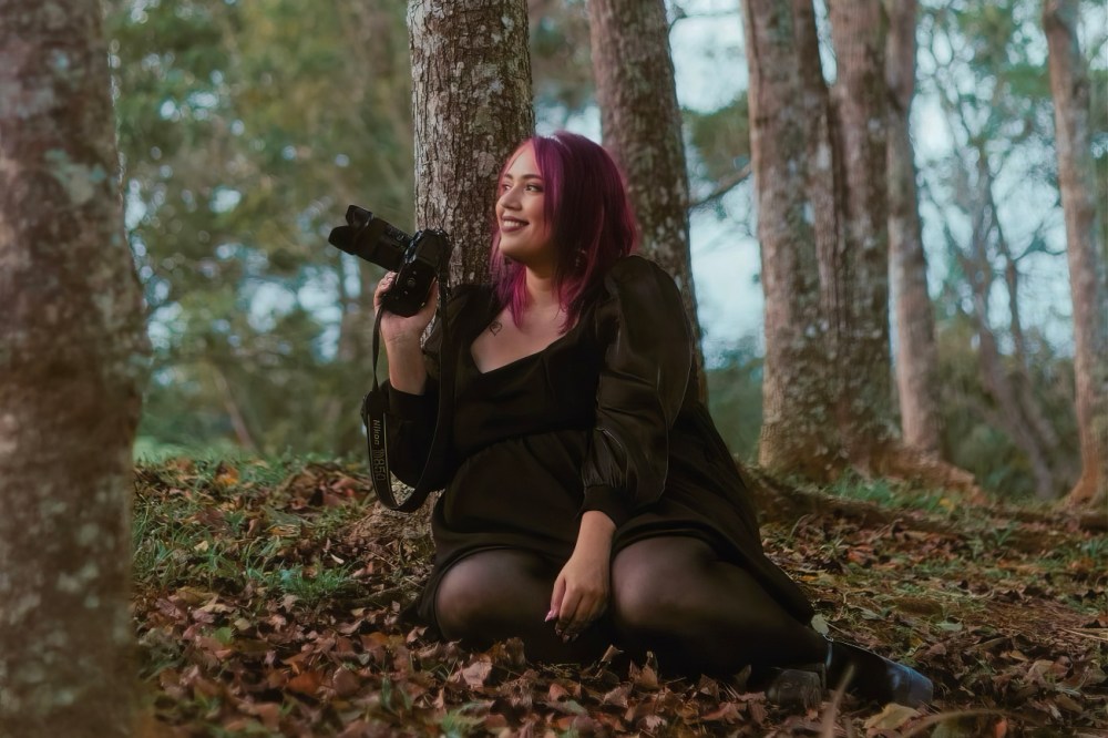 Person with purple hair holding camera, sitting on forest ground.