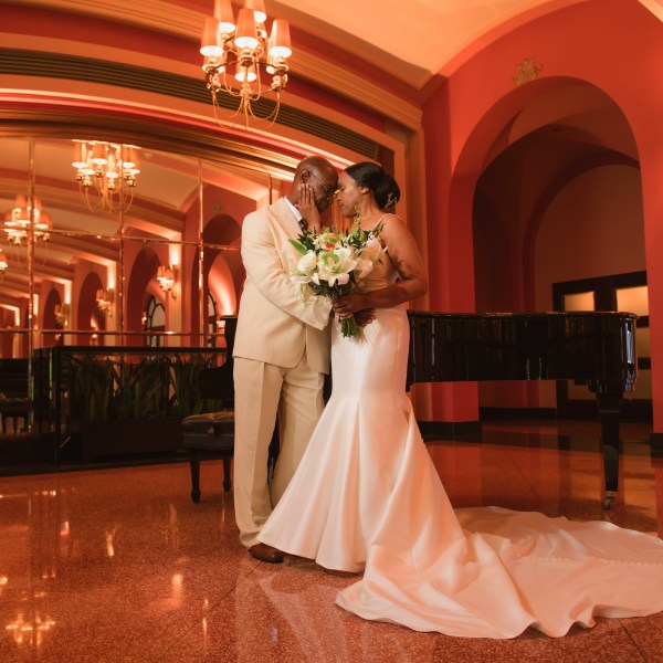 Couple in formal attire embraces in elegant room with mirrors and chandelier.